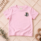 I AM Kids Crowned JC Jesus Christ Kids Tee | Left Chest design on a Light Pink shirt, shown on a cream rug.