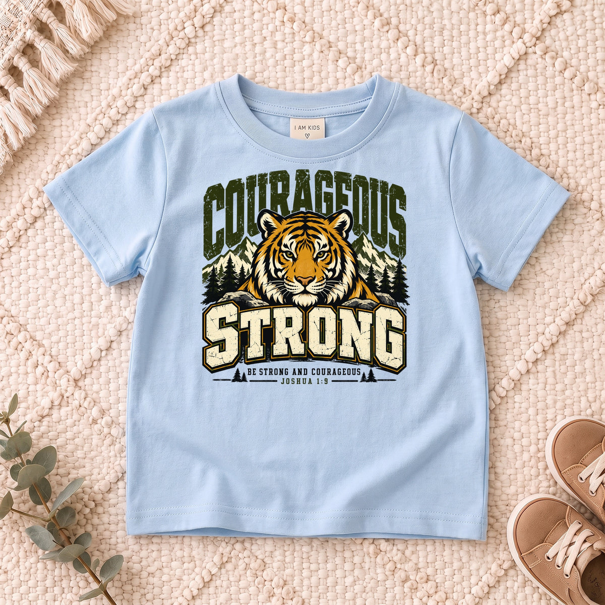 I AM Kids Be Strong & Courageous Tiger Kids Tee design on a Light Blue shirt, shown on a cream rug.