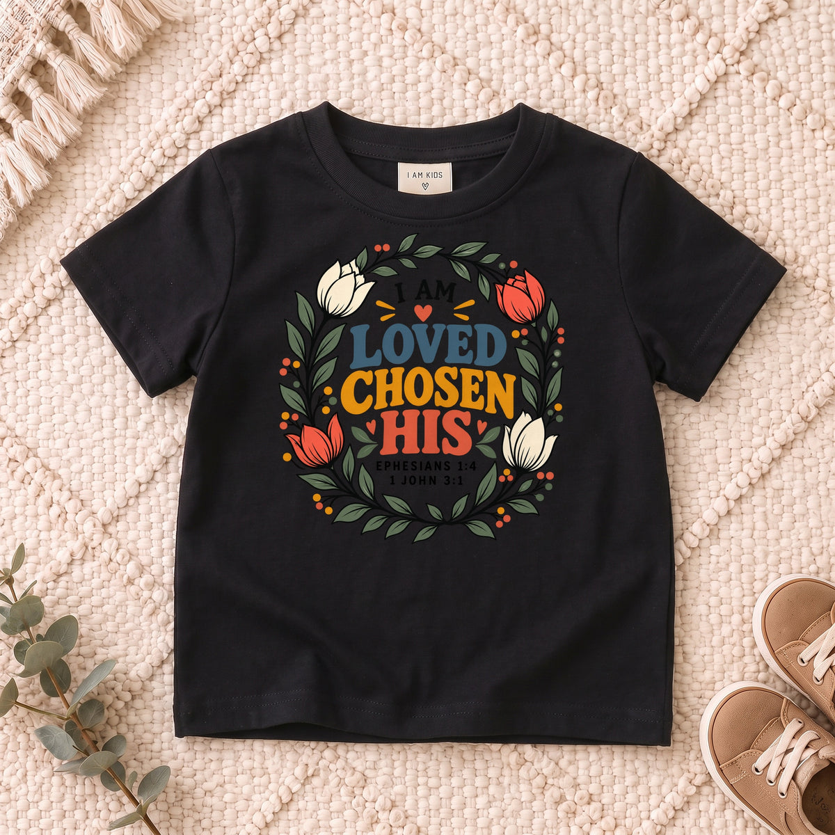 I AM Kids I AM Loved, Chosen & His Floral Wreath Kids Tee design on a Black shirt, shown on a cream rug.