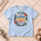 I AM Kids I AM Loved, Chosen & His Floral Wreath Kids Tee design on a Light Blue shirt, shown on a cream rug.
