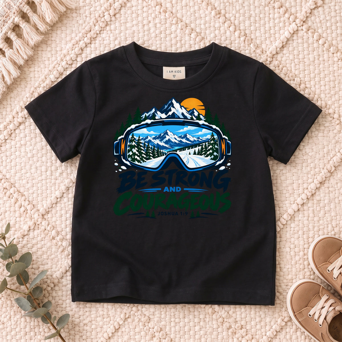 I AM Kids Be Strong & Courageous Mountain Goggles Kids Tee | Joshua 1:9 design on a Black shirt, shown on a cream rug.