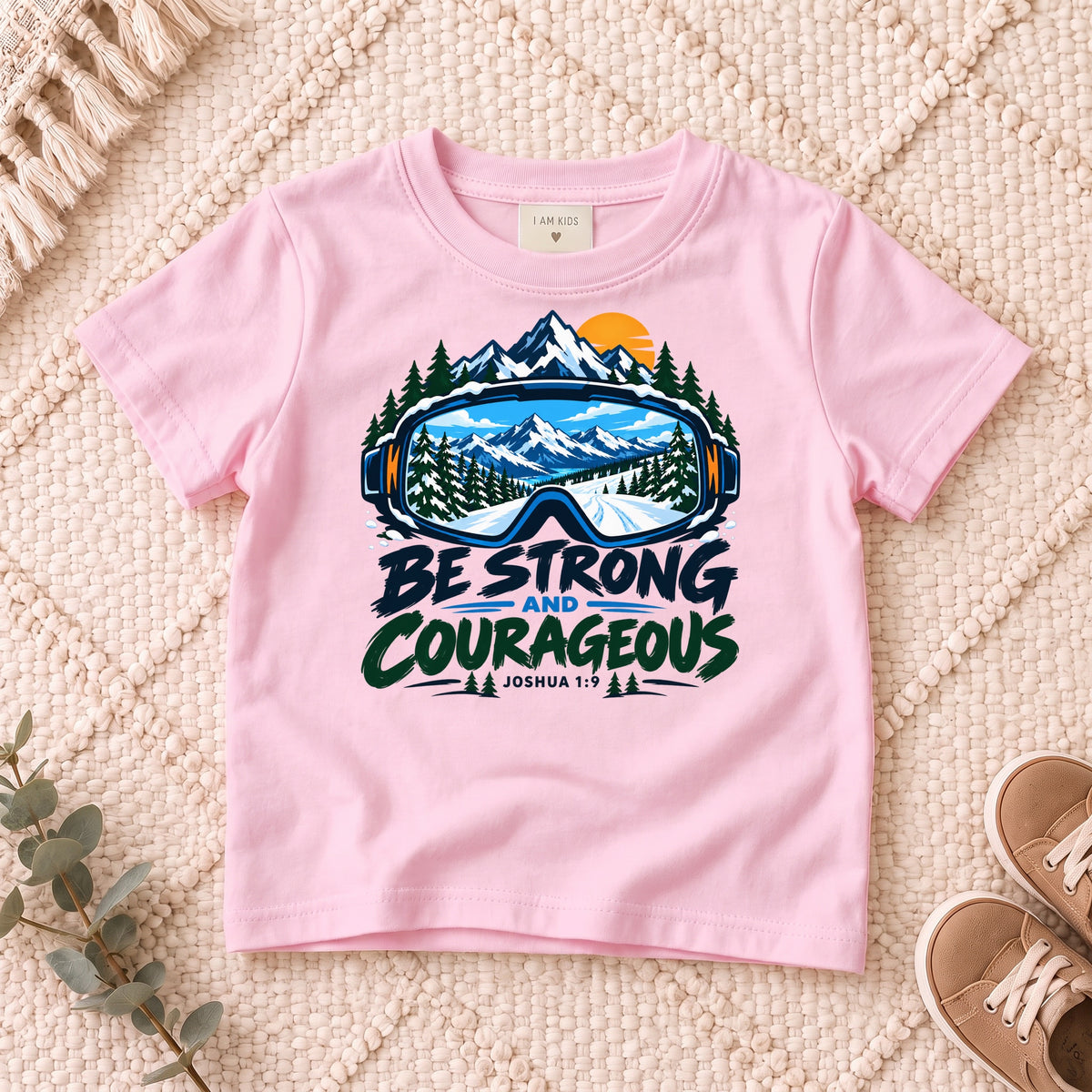 I AM Kids Be Strong & Courageous Mountain Goggles Kids Tee | Joshua 1:9 design on a Light Pink shirt, shown on a cream rug.