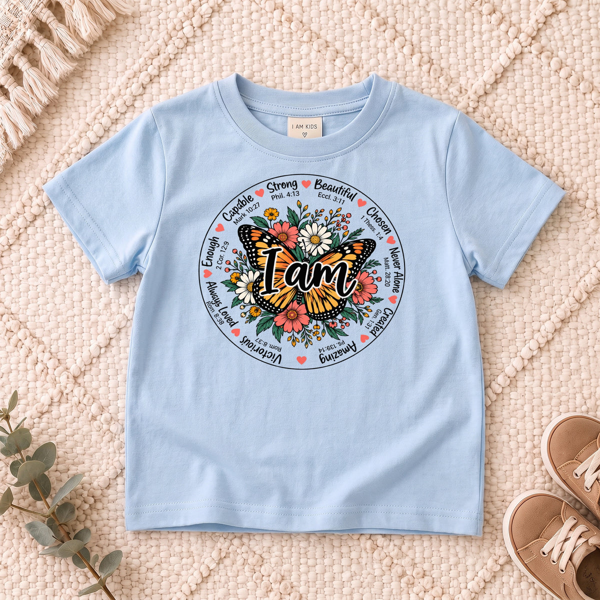 I AM Kids I AM Butterfly Words of Faith Kids Tee design on a Light Blue shirt, shown on a cream rug.